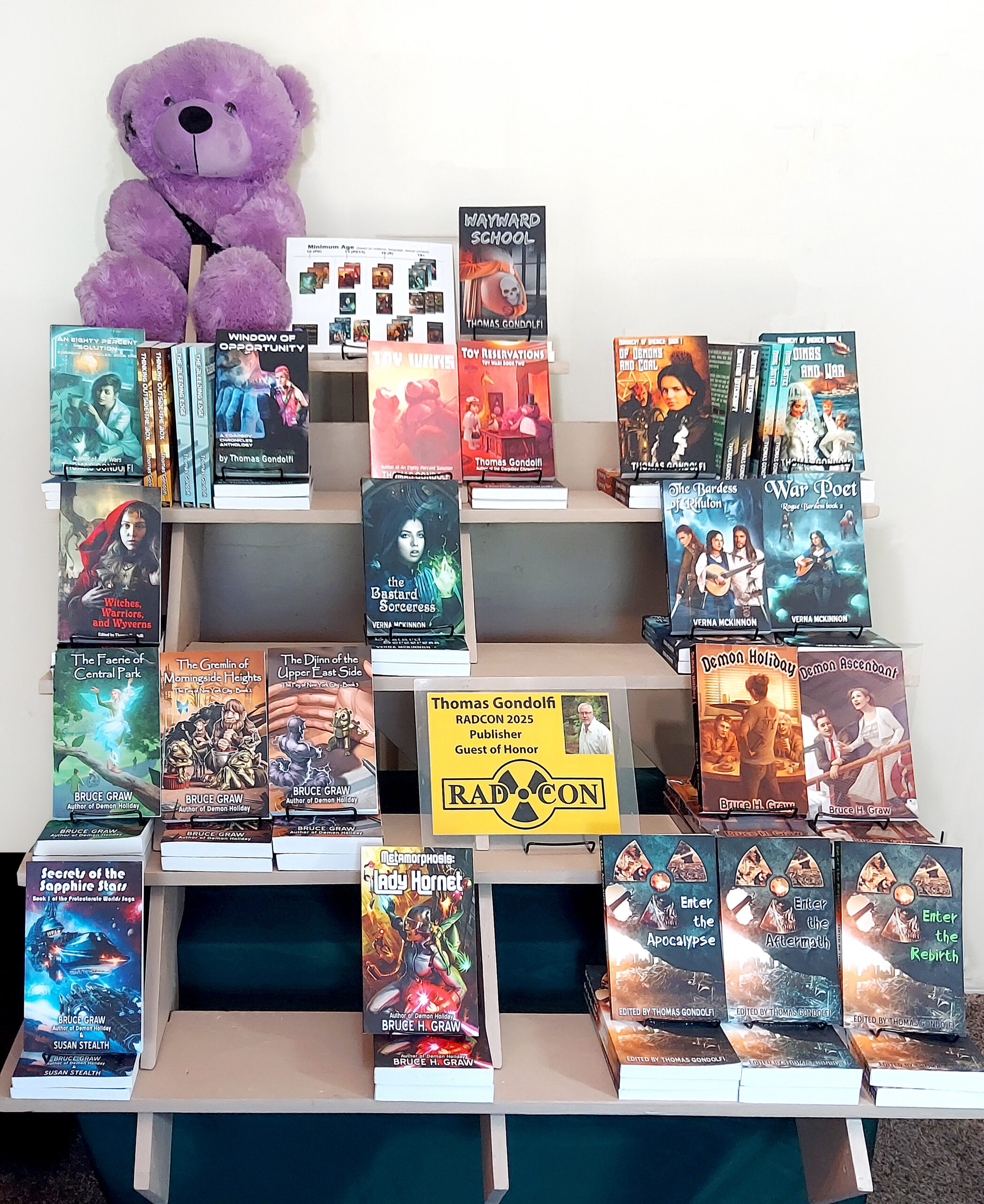 A display at a book fair or convention showcasing a variety of books. In the center, a large purple teddy bear sits atop a table with multiple shelves filled with books. The books are arranged by author, with titles such as Wayward School by Thomas Gondolfi, The Bastard Sorceress by Verna McKinnon, and Demon Ascendant by Bruce H. Graw. In front of the display is a sign reading "Thomas Gondolfi, RADCON 2025 Publisher, Guest of Honor.